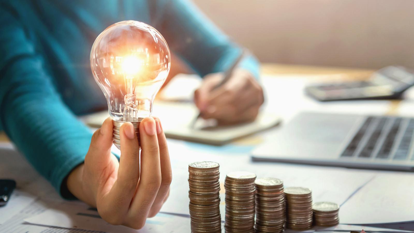 Woman Calculating Finances and Holding Lightbulb with Stack of Coins on Table